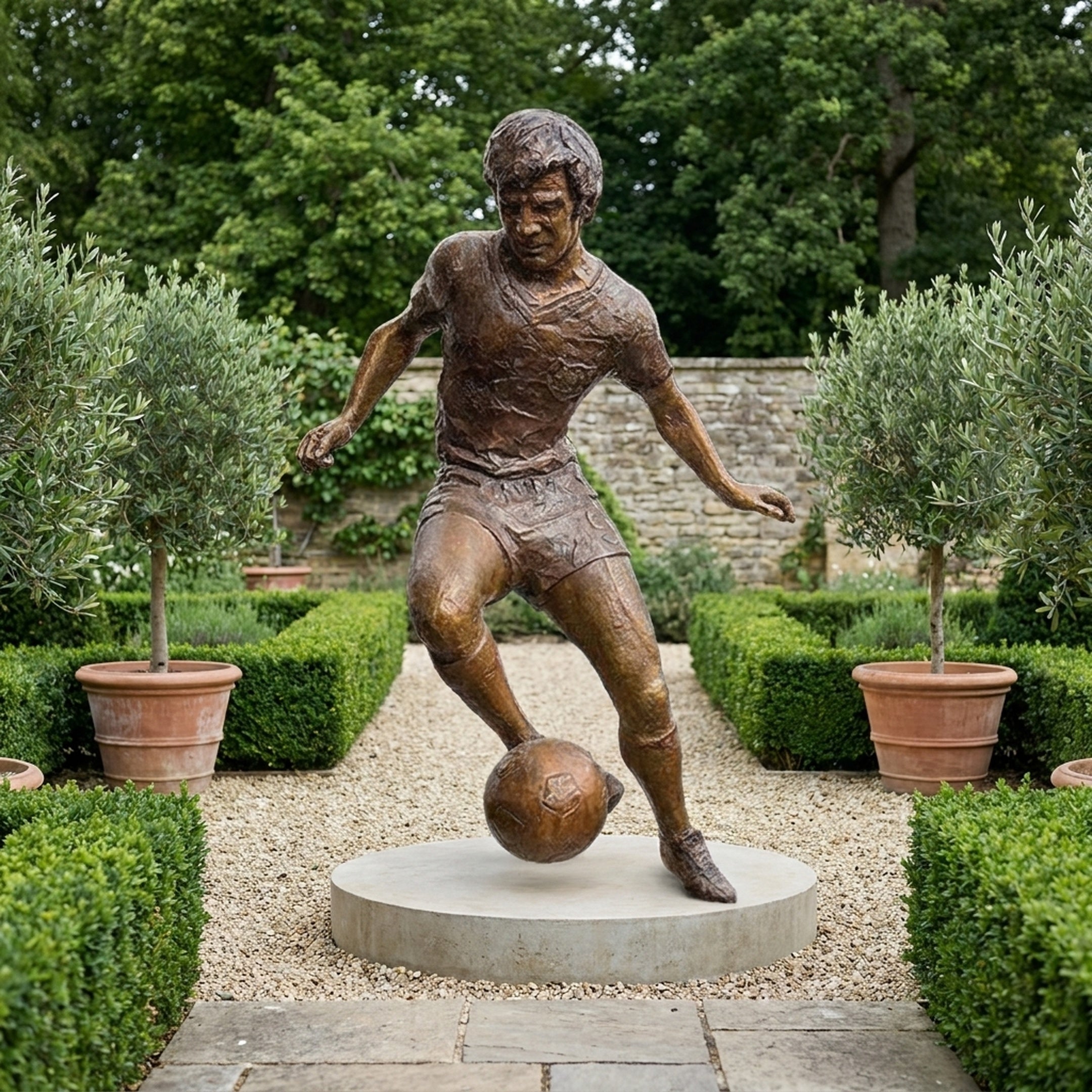 Life-Size Contemporary Footballer Dribbling Bronze Sculpture by Giant Sculptures shown mid-stride with the ball on a round stone base in a hedged garden.