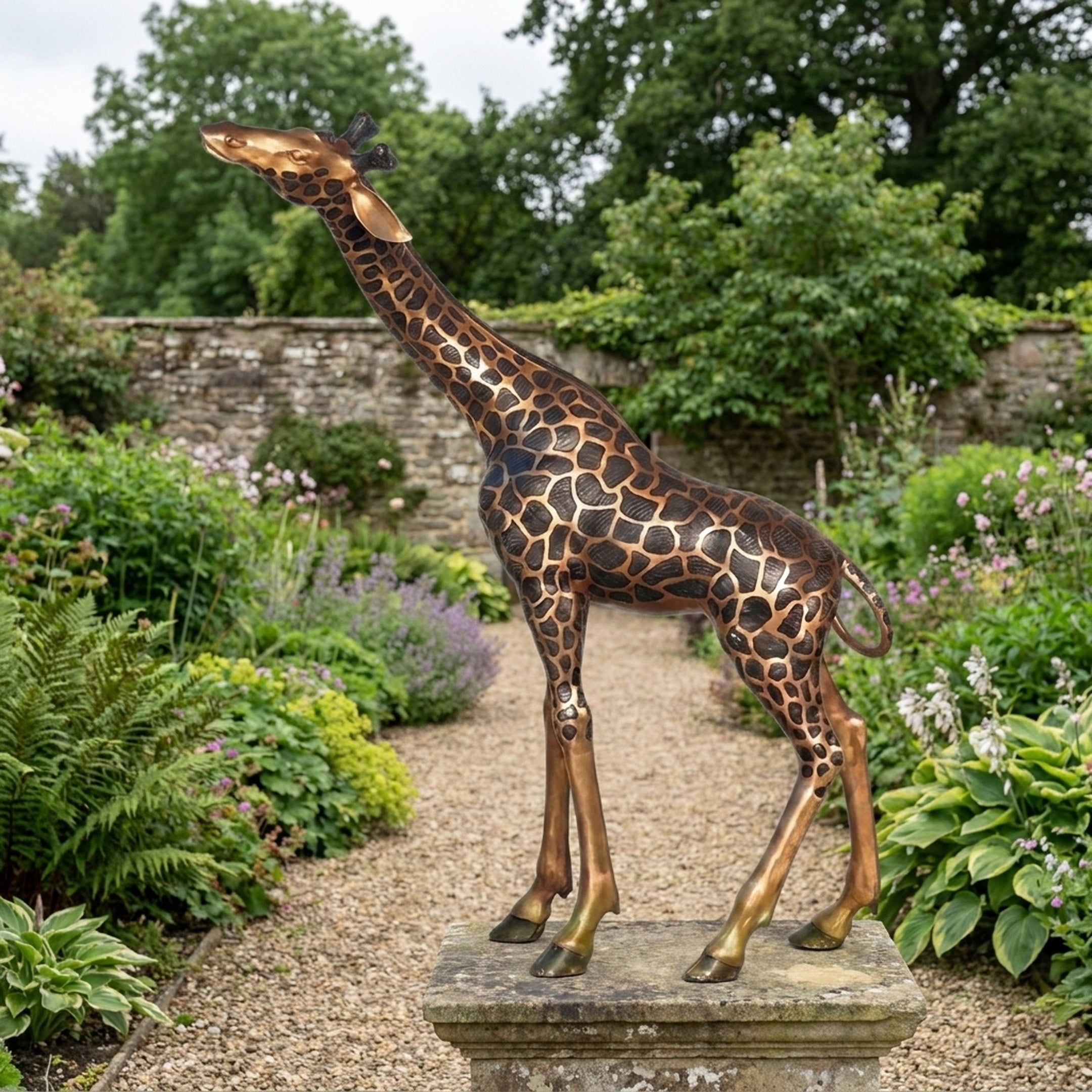 Life-Size Contemporary Giraffe Bronze Sculpture by Giant Sculptures with two-tone patina, displayed on a stone plinth along a gravel garden path with lush borders.