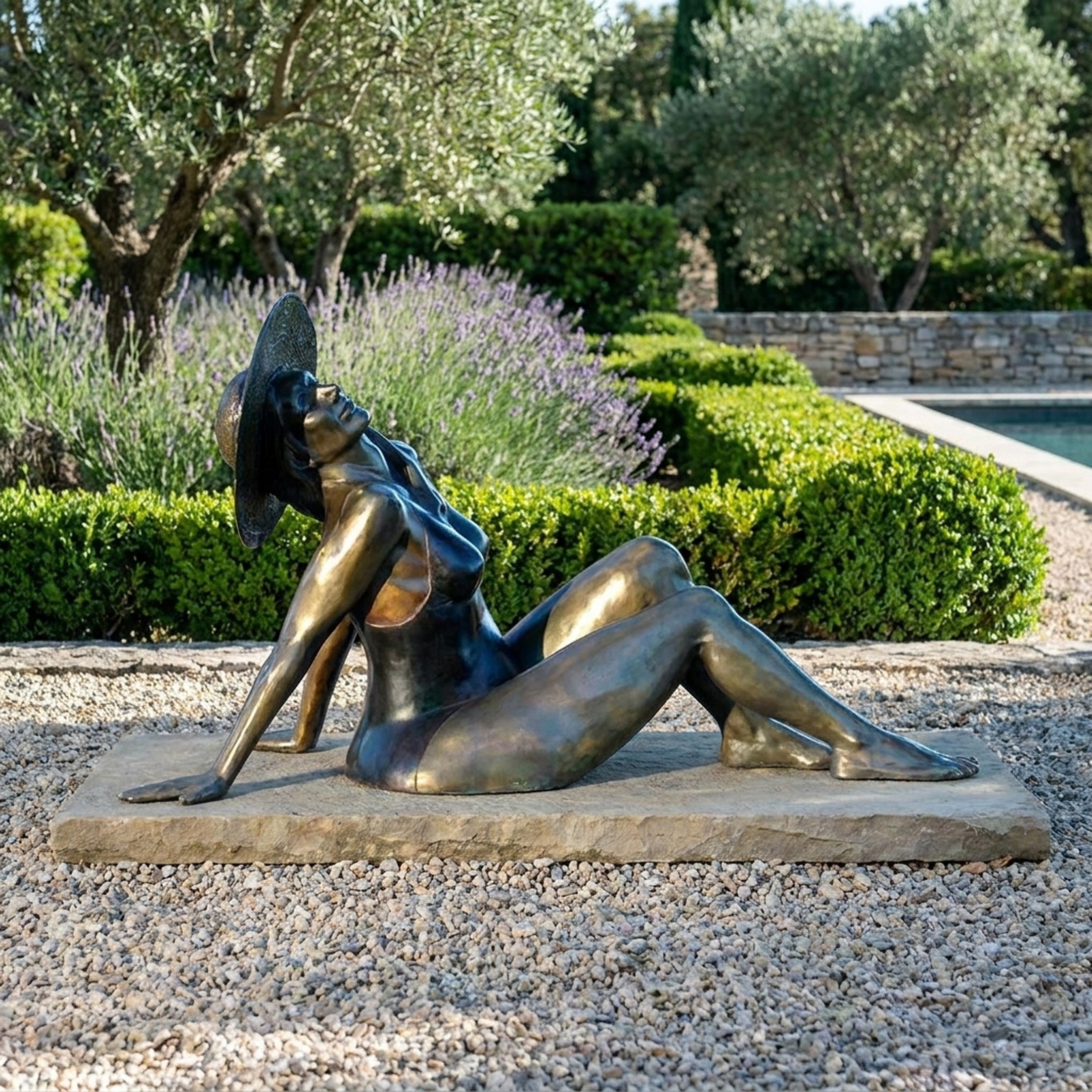 Giant Sculptures Life-Size Contemporary Reclining Woman Bronze Sculpture with wide-brimmed hat on a stone slab in a lavender garden by a pool.