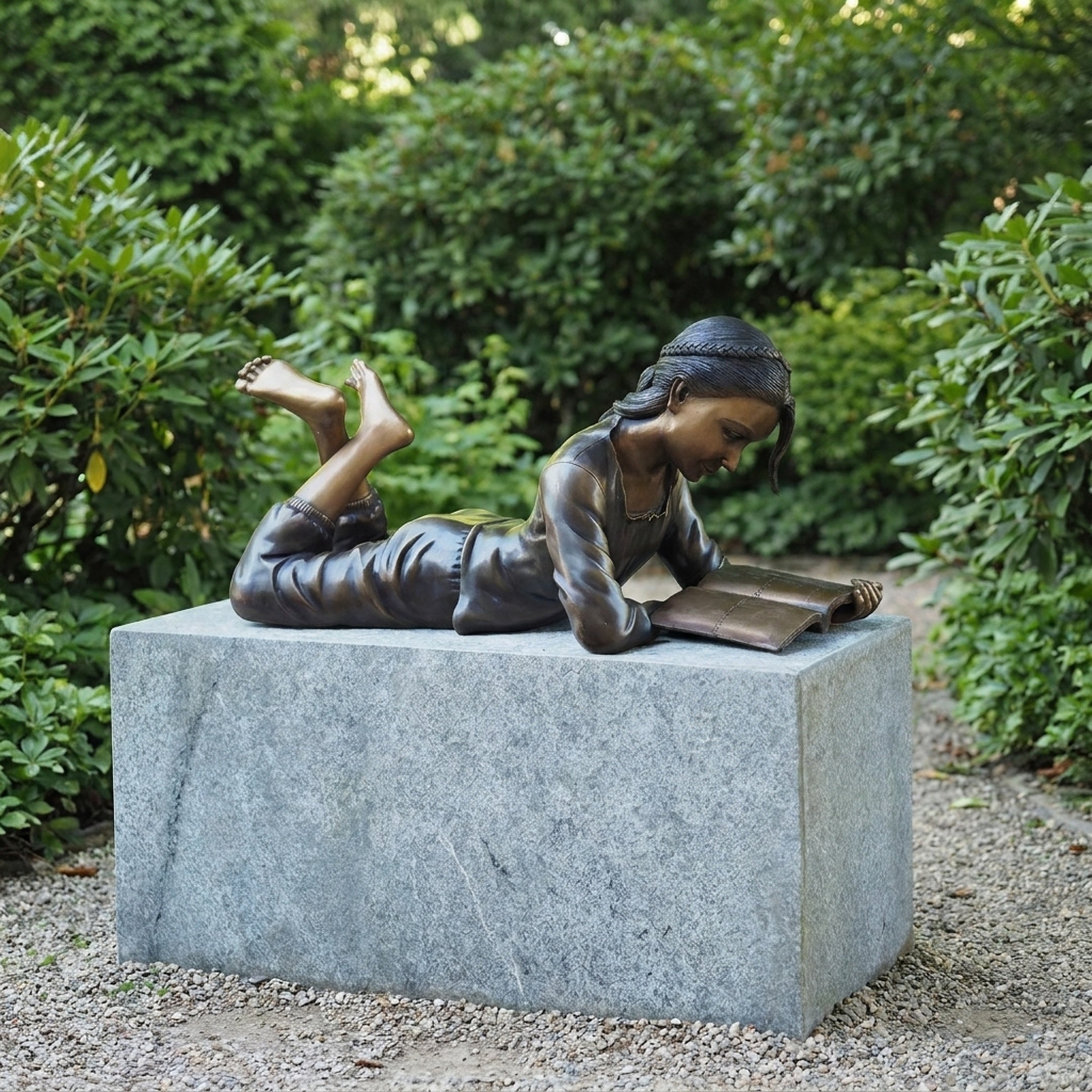 Life-Size Girl Reading Book Bronze Sculpture by Giant Sculptures, shown lying prone on a granite plinth in a garden setting with green shrubs behind.