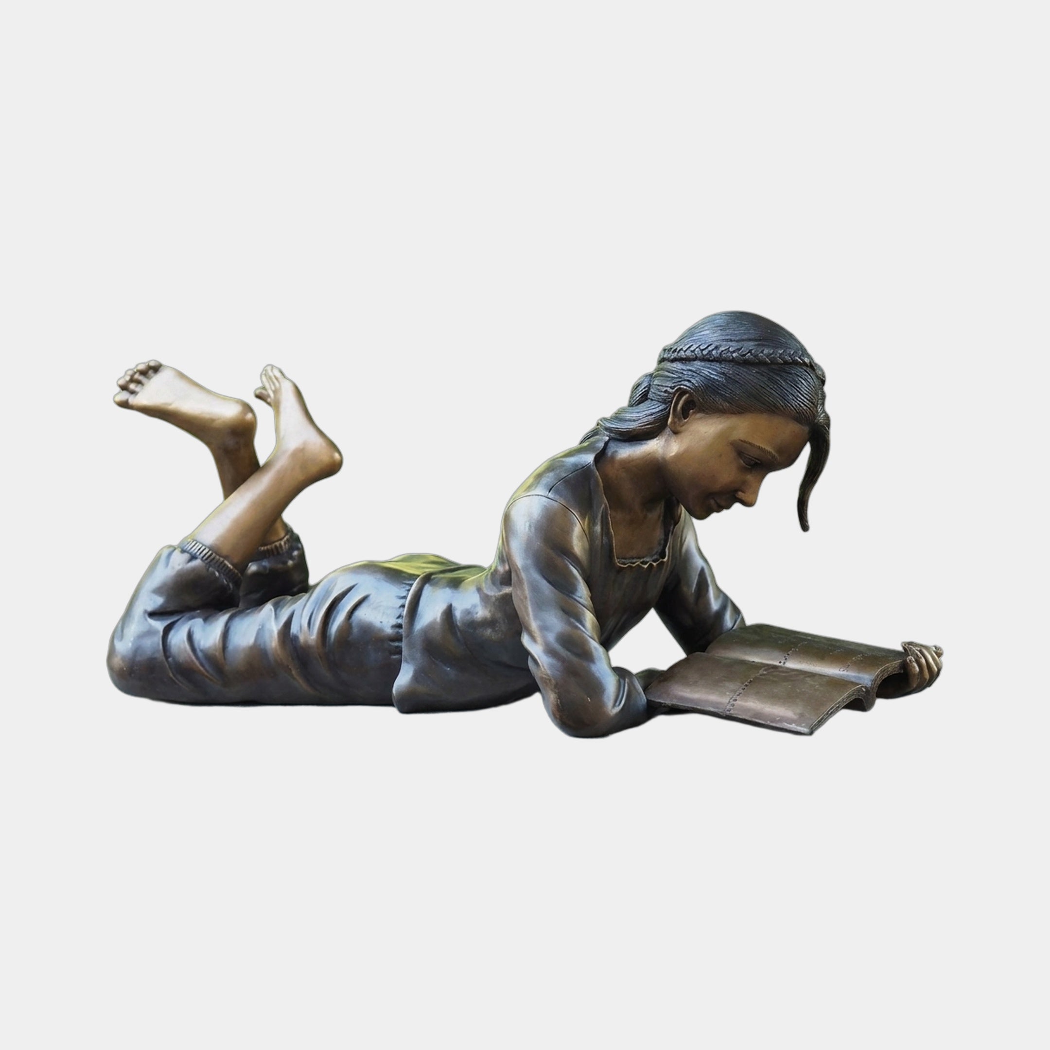 Life-Size Girl Reading Book Bronze Sculpture by Giant Sculptures, shown lying prone with bare feet raised and head bent over an open book, on a plain background.