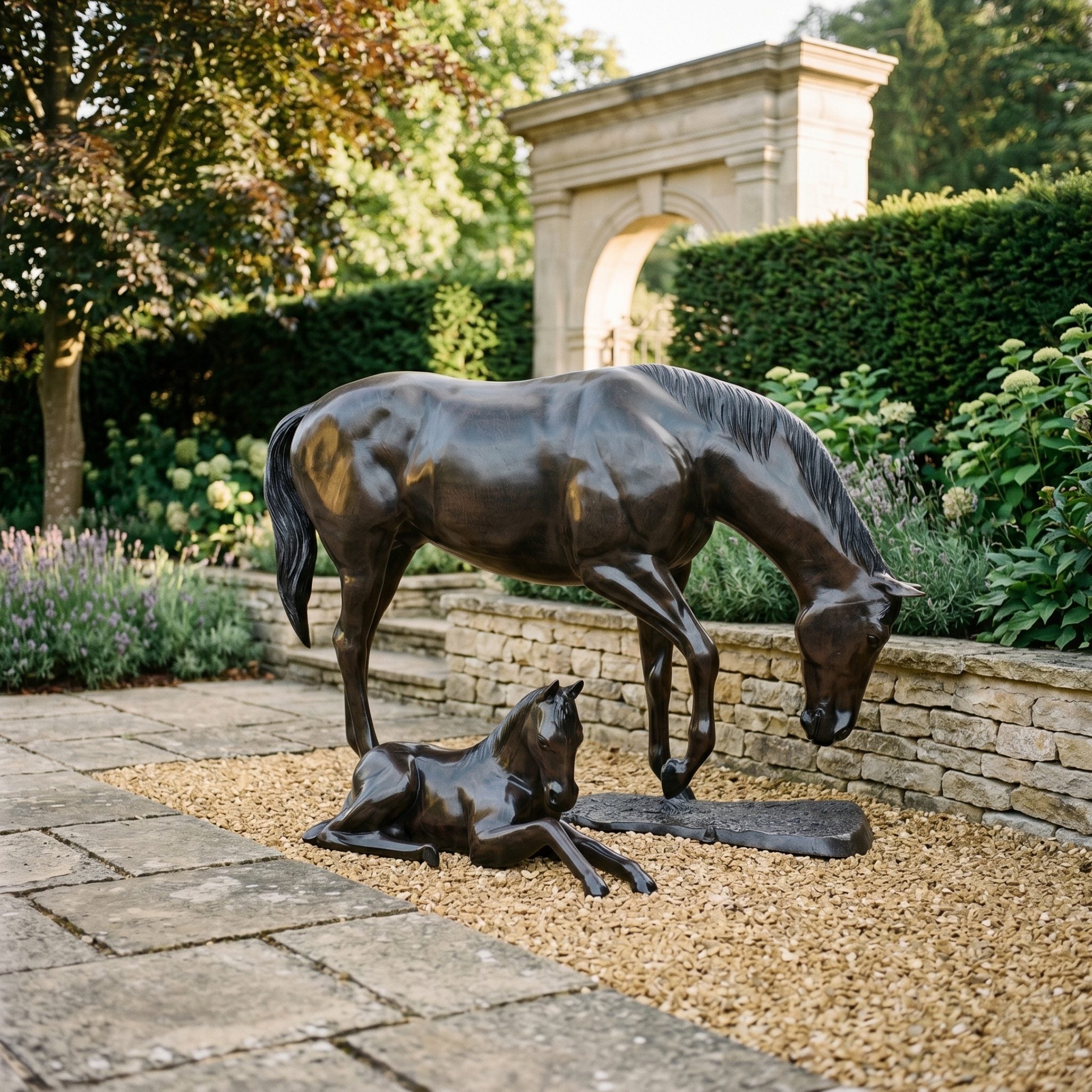Life-Size Mare and Foal Bronze Sculpture by Giant Sculptures, dark patinated mare grazing beside recumbent foal on gravel in a stone garden with arched gateway.