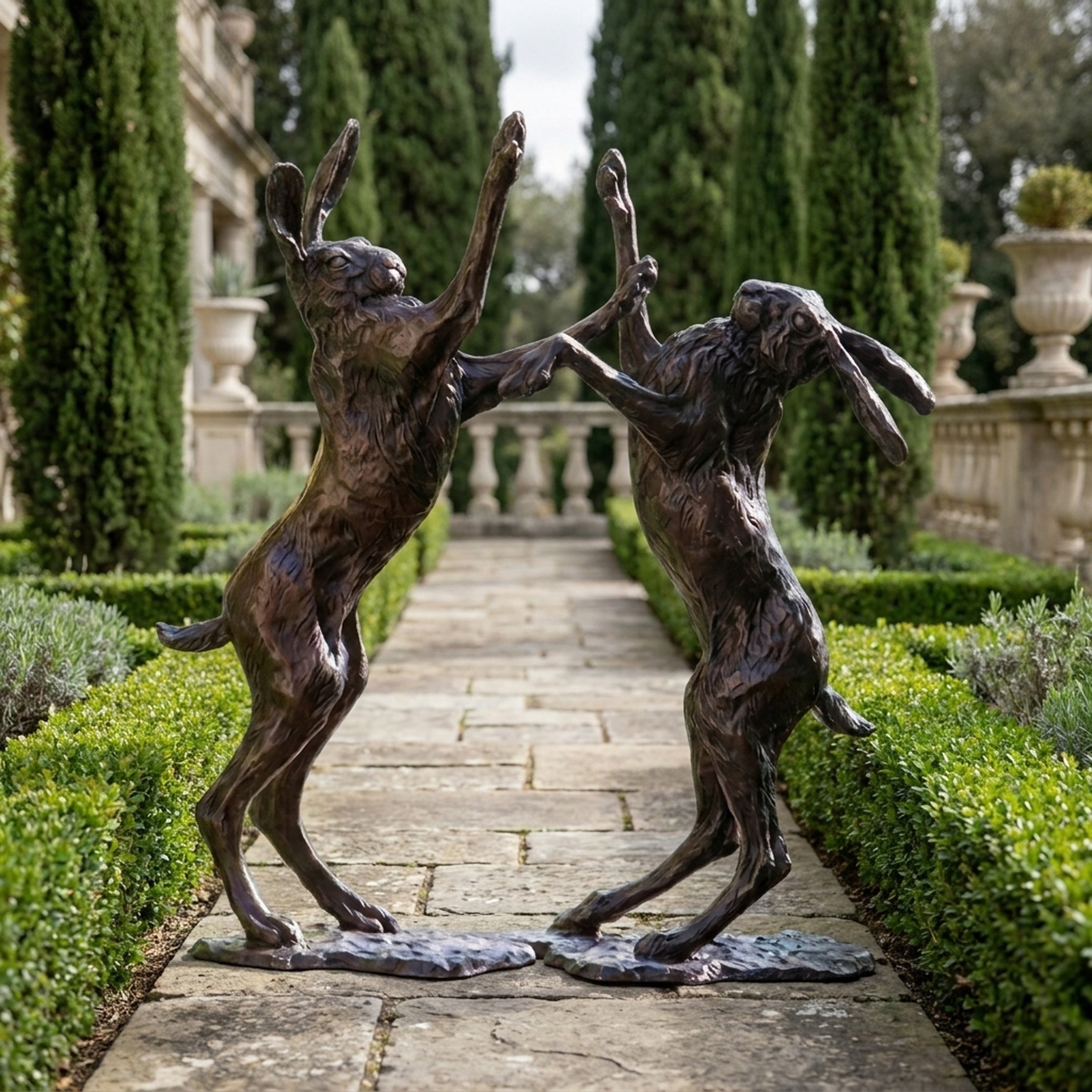 Life-Size Traditional Boxing Hares Bronze Sculpture by Giant Sculptures, two dark patinated hares rearing on hind legs on a garden path lined with cypress trees.