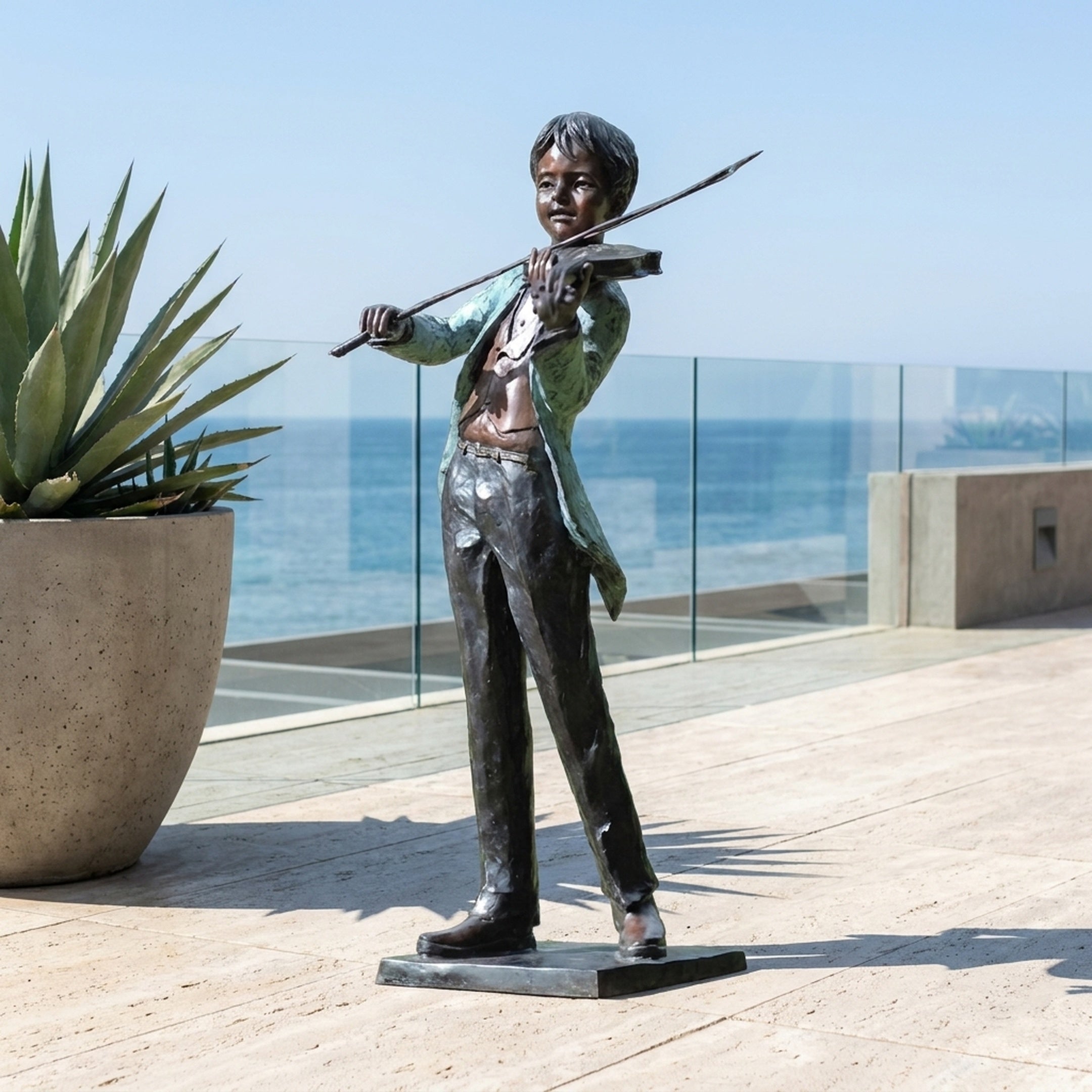 Life-Size Traditional Boy Violinist Bronze Statue by Giant Sculptures with dark patina and green verdigris jacket, displayed on a seaside terrace beside an agave planter.