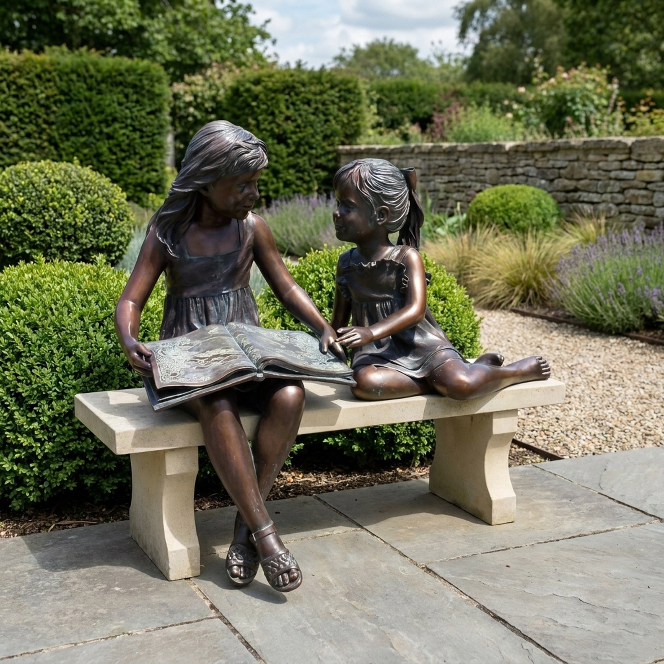 Life-Size Two Girls Reading on a Bench Bronze Sculpture by Giant Sculptures, 130cm, displayed on a stone patio beside lavender and clipped hedges.