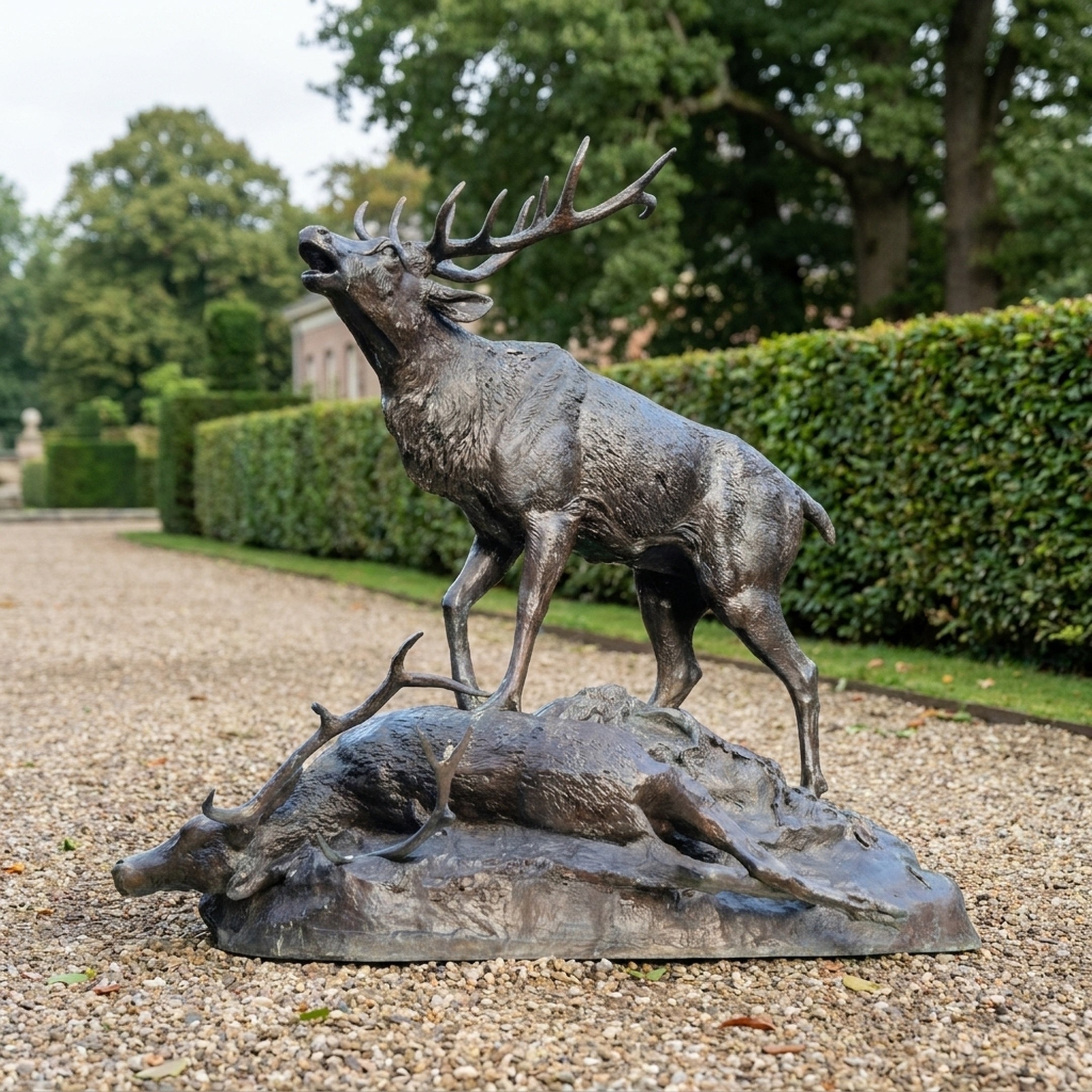 Monumental Bugling Elk and Fallen Deer Bronze Sculpture by Giant Sculptures, dark brown patina, shown on a gravel garden path beside trimmed hedges.