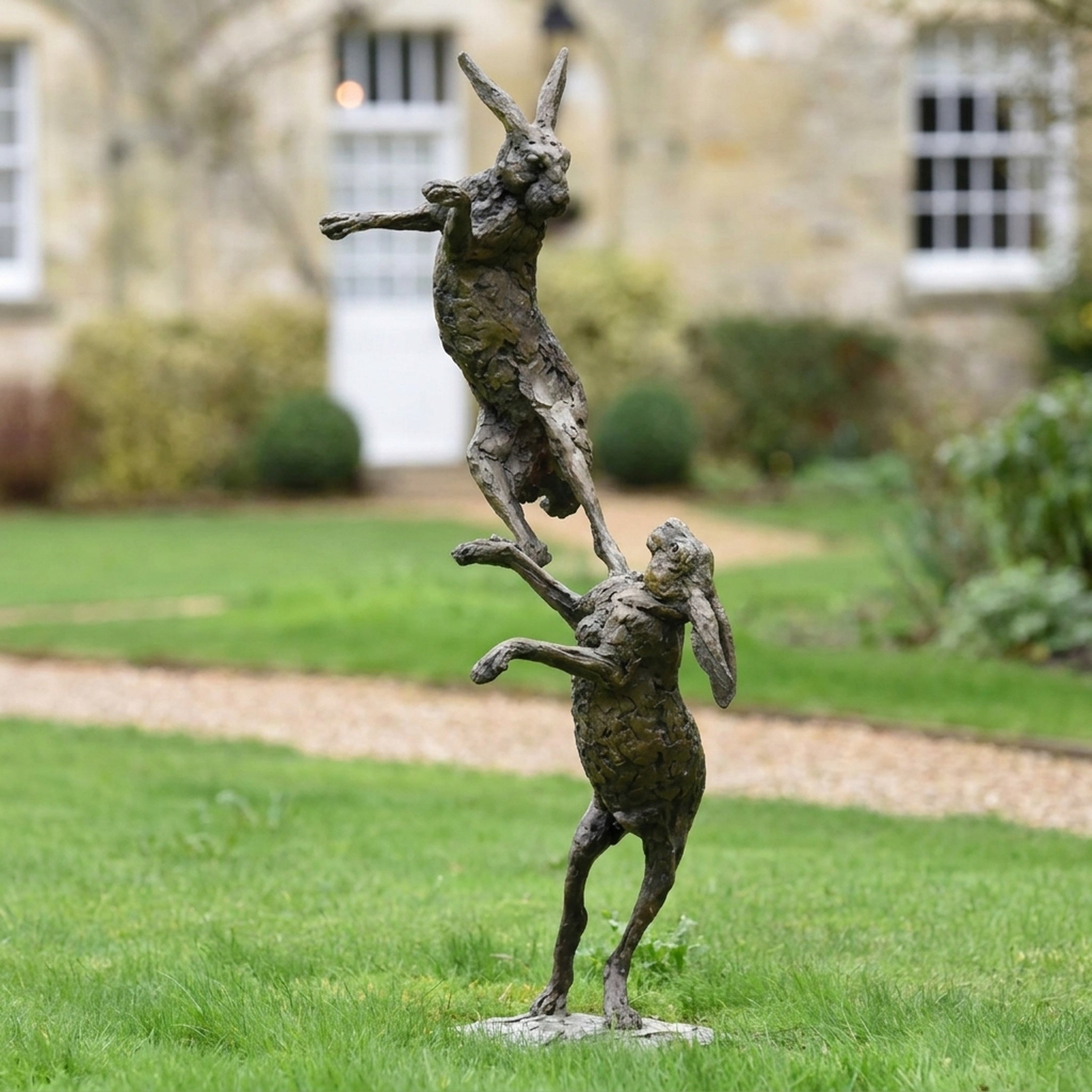 Monumental Contemporary Boxing Hares Bronze Sculpture by Giant Sculptures, two stacked hares with dark patina displayed on a lawn before a stone house.