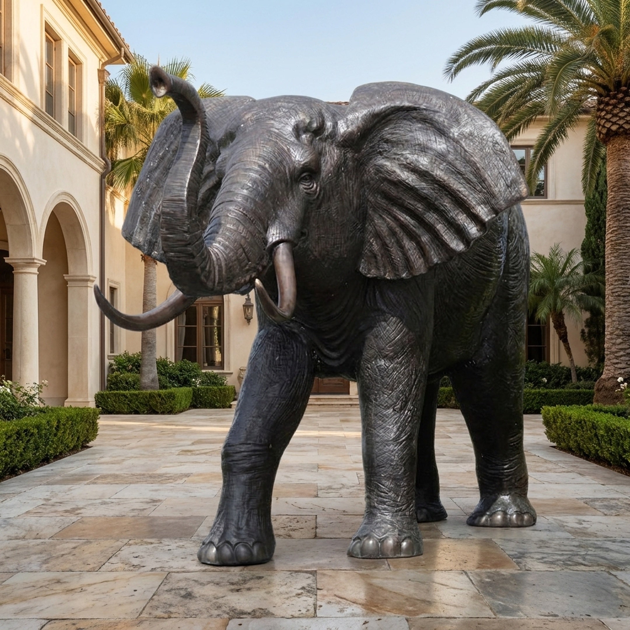Monumental Traditional African Elephant Bronze Sculpture by Giant Sculptures with raised trunk and pale tusks, displayed on a paved courtyard among palms.