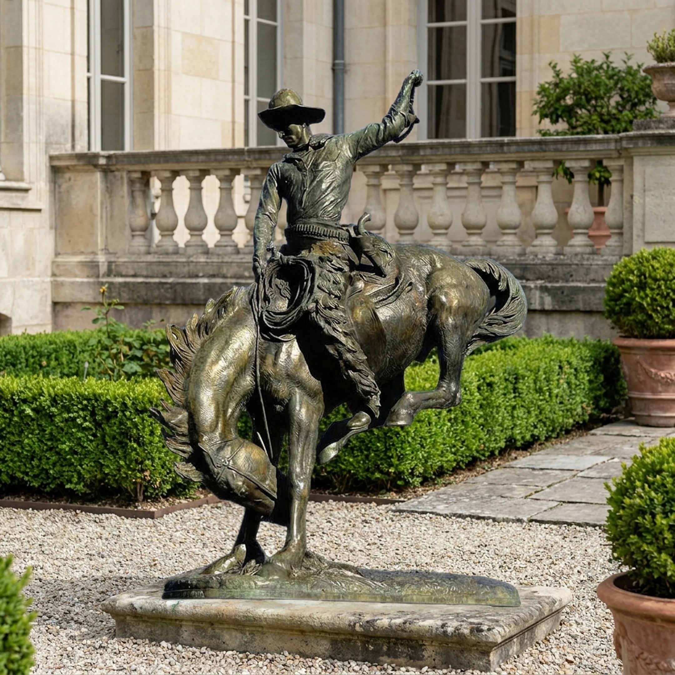 Monumental Traditional Cowboy and Bucking Bronco Bronze Sculpture by Giant Sculptures, rider raising hat as the horse rears, displayed on gravel by a stone balustrade.