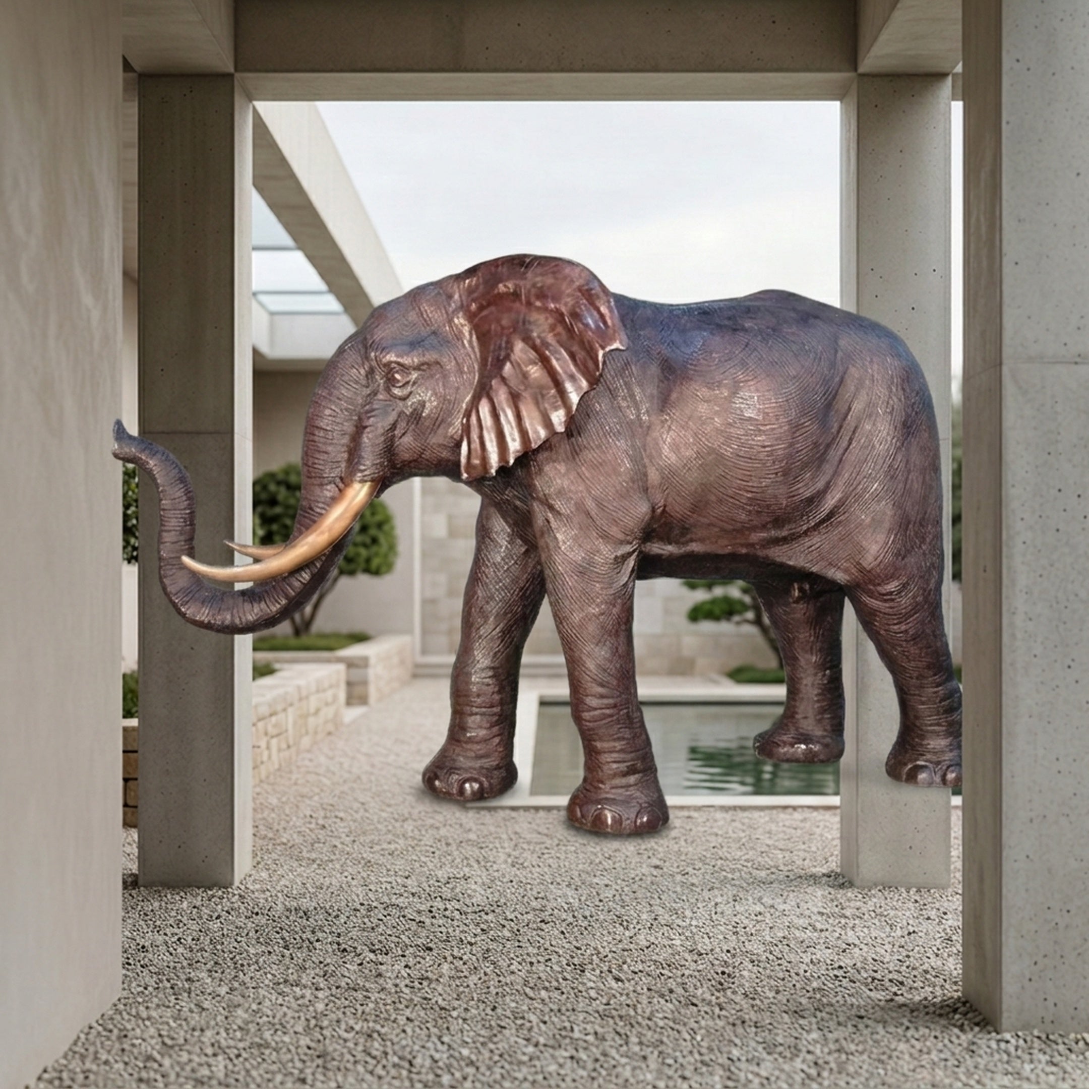 Monumental Traditional Walking Elephant Bronze Sculpture by Giant Sculptures shown in profile with raised trunk and ivory tusks in a concrete courtyard.