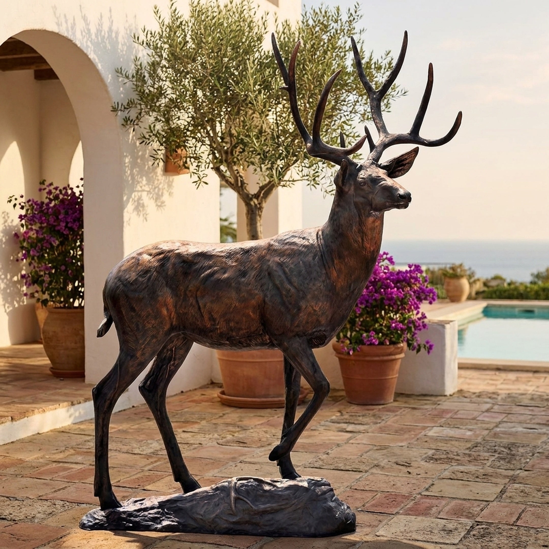 Monumental Traditional Walking Stag Bronze Sculpture by Giant Sculptures, 280cm, shown mid-stride on a rocky base in a sunlit Mediterranean poolside courtyard.