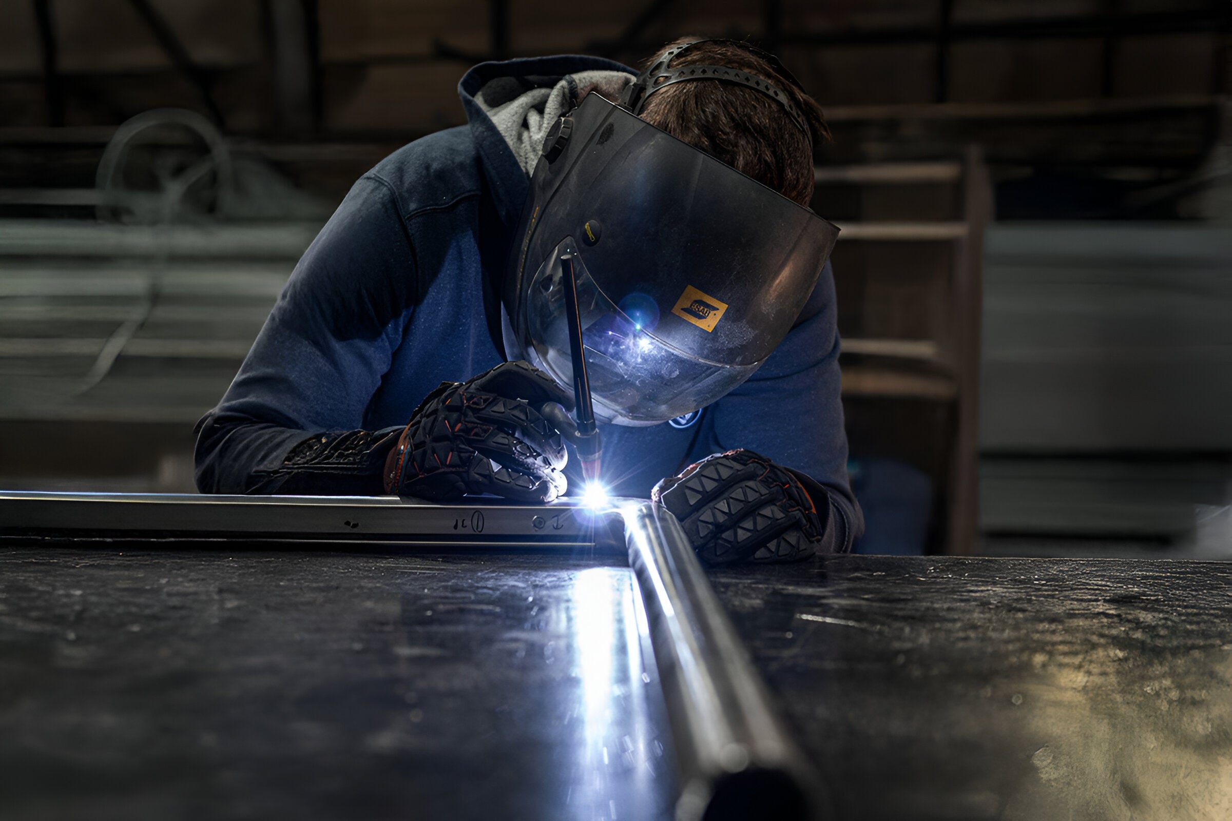 A welder wearing a dark visor helmet and gloves works intently on a metal pipe. Bright sparks and light are visible at the welding point, set against a dimly lit workshop background.
