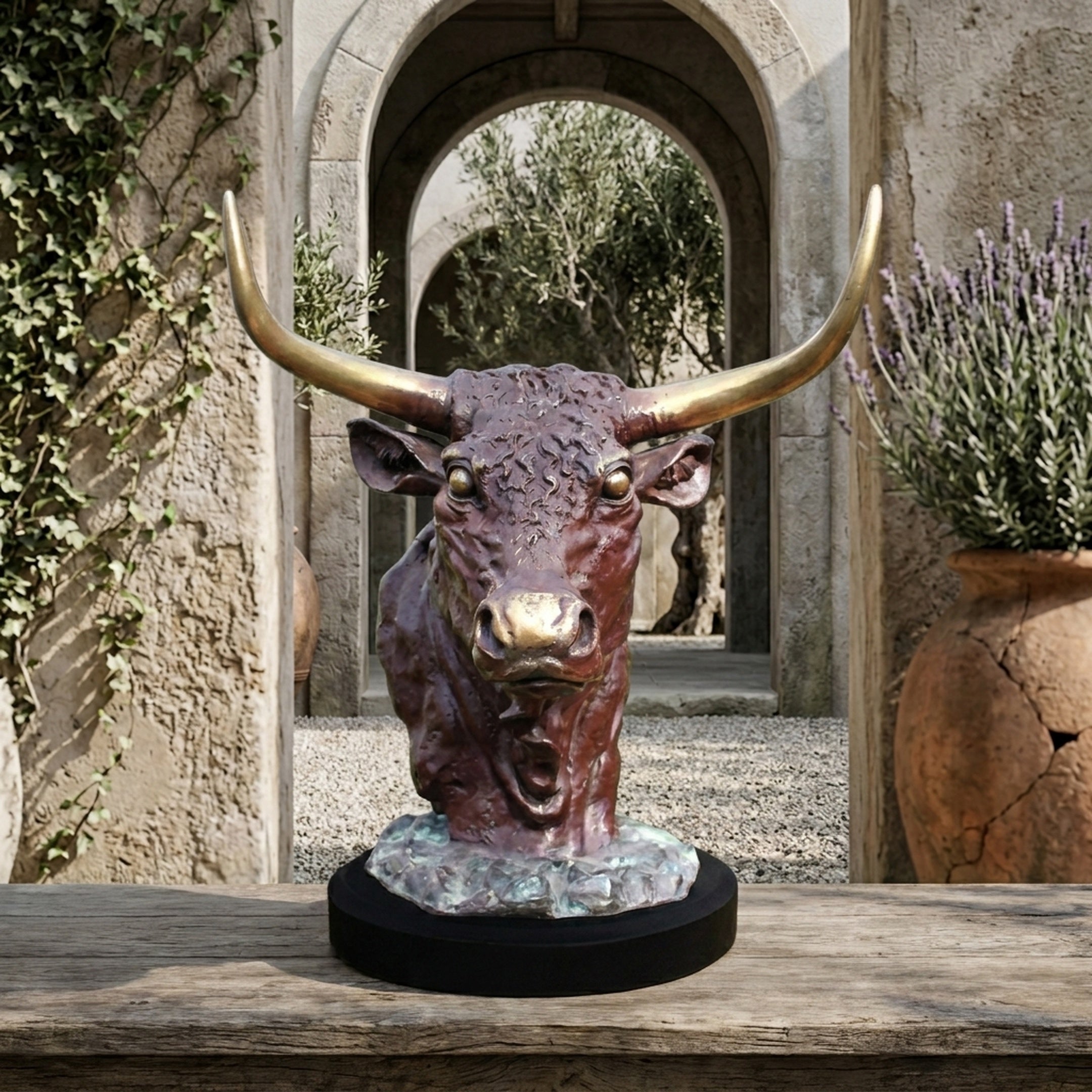 Traditional Bull Head Bronze Bust by Giant Sculptures with reddish-brown patina and golden horns on a black plinth, displayed outdoors near a stone archway.