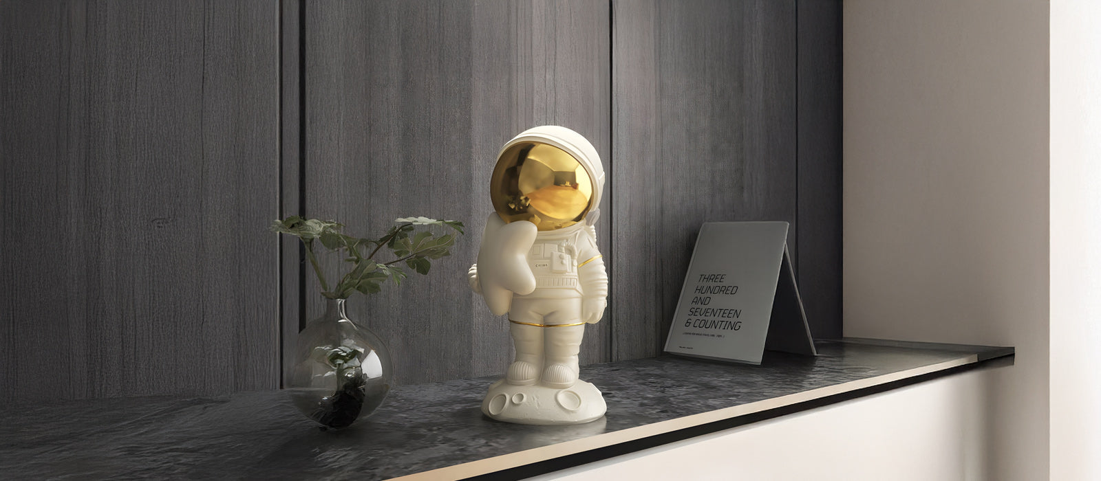 A small astronaut figurine with a gold visor stands on a black countertop next to a modern vase with a green plant and a tilted rectangular sign with text, in a room with dark wooden walls.