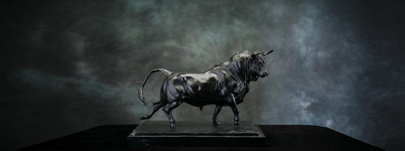Dark bronze-finish bull sculpture in a powerful mid-stride pose on a rectangular marble-look base, set against a moody grey-green studio backdrop.