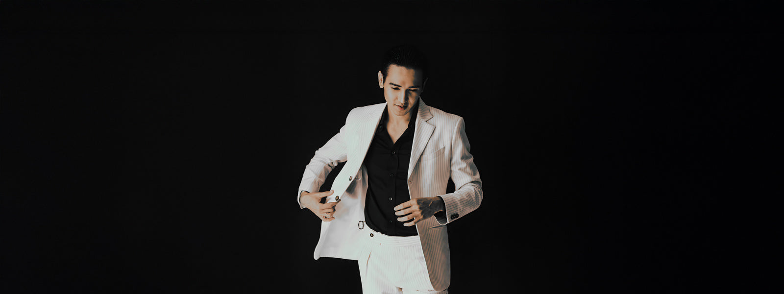 A man in a white suit stands against a dark background, looking down with a slight smile. He is adjusting his suit jacket with one hand.