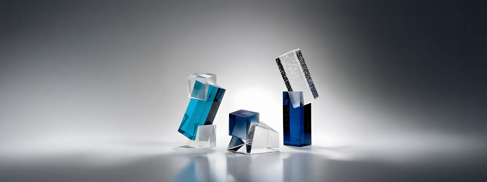 Three modern glass sculptures with geometric shapes in clear, blue, and frosted glass stand on a smooth surface, dramatically lit against a gradient grey background.