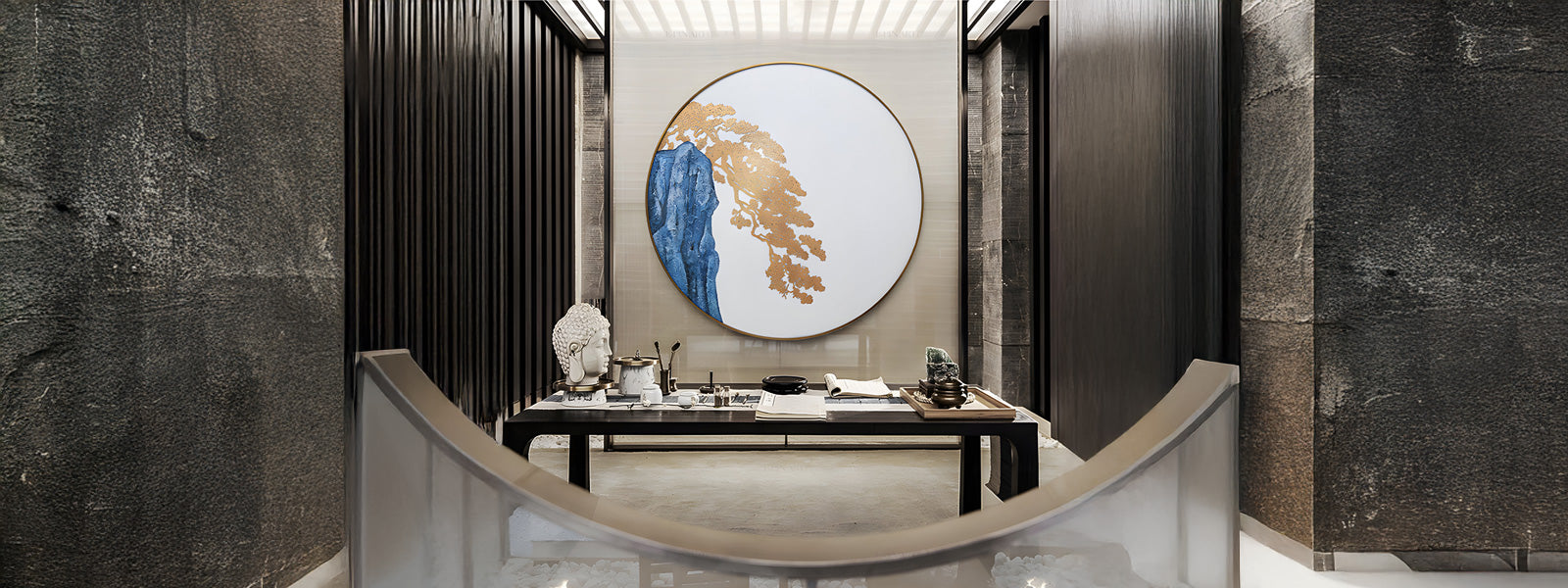 A modern interior with a round, abstract art piece featuring gold and blue colors on the wall. Below is a sleek dark table displaying decorative items, flanked by tall, textured walls.