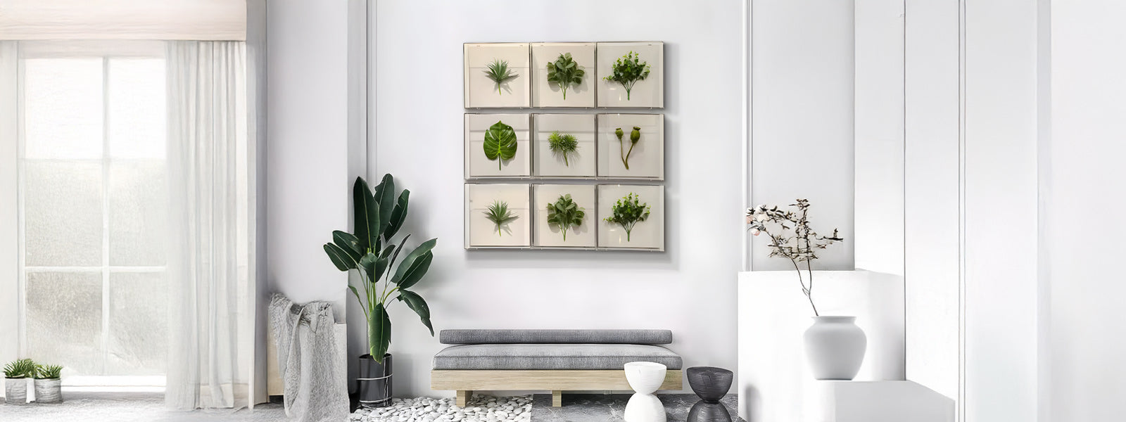 A minimalist living room with white walls, a large window, and a plant in a corner. Artwork featuring framed plants hangs on the wall above a gray bench. A vase with branches sits on a white cabinet.