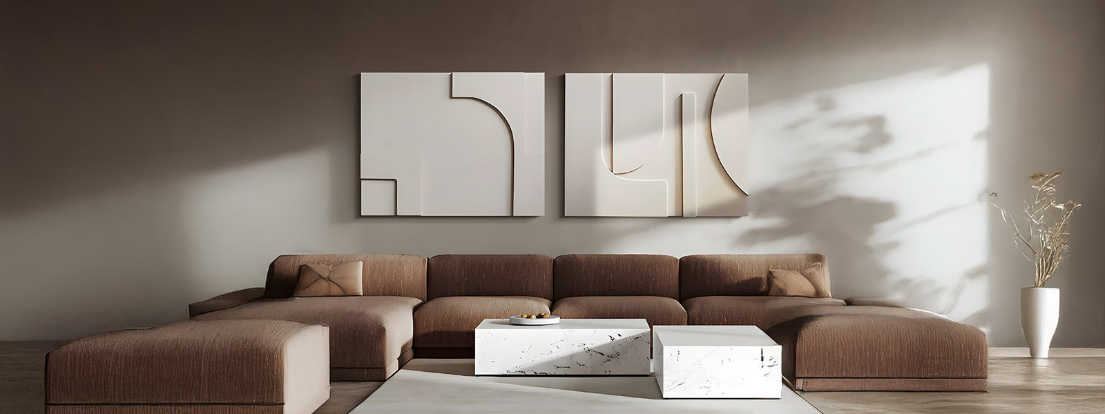 A modern living room features a large sectional brown sofa, abstract wall art, a white marble coffee table, and a white vase with dried branches. Sunlight casts shadows on the neutral-toned wall.