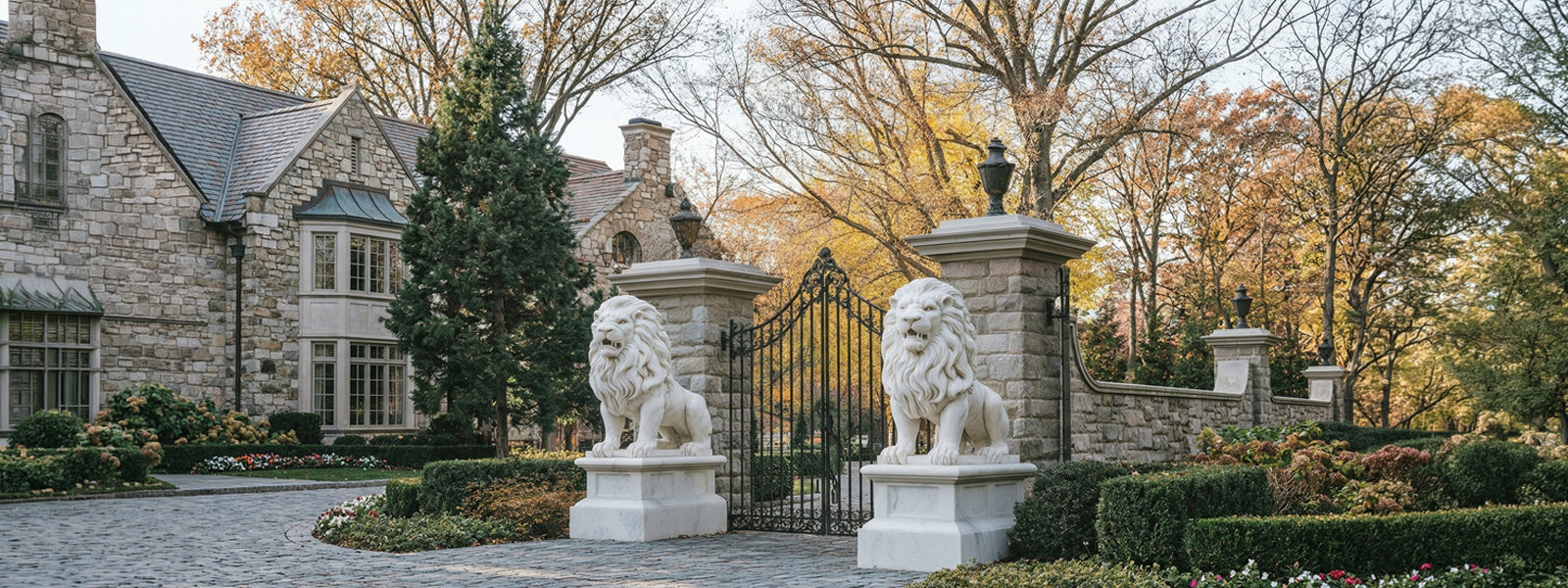 Pair of white lion statues on stone pedestals flanking a wrought-iron gate at the entrance of a stone manor house surrounded by autumn foliage.