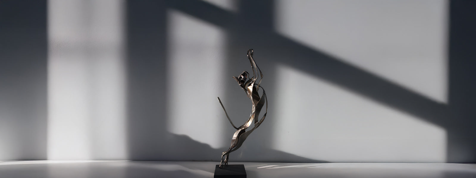 A sleek metal sculpture of a leaping cat is displayed on a flat surface, with dramatic shadows and light patterns cast on the plain wall behind it.