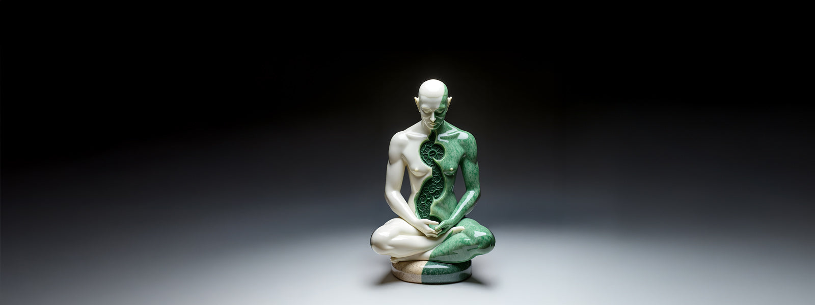 A sculpture of a bald, nude human figure sitting cross-legged, with one half smooth and white, the other half textured and green, split down the center. The background is dark and minimal.