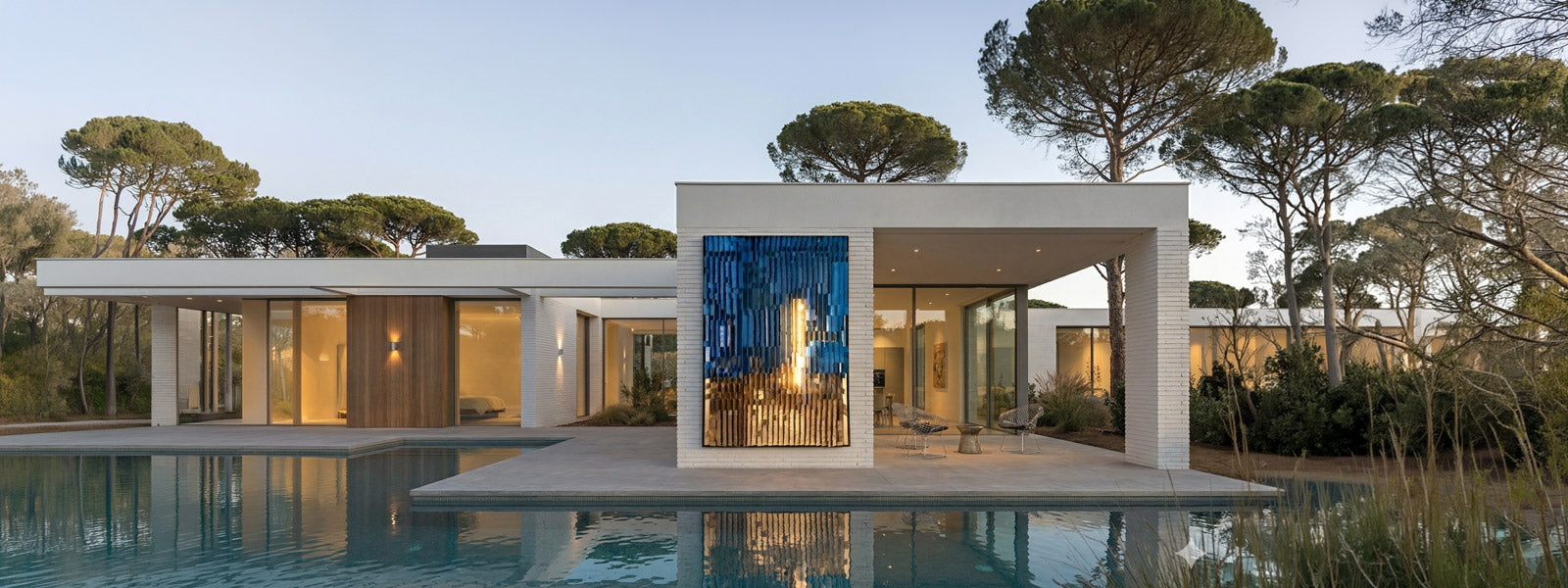 Modern white villa with tall blue and gold outdoor wall art panel between brick columns, reflective pool and Mediterranean pines at dusk.