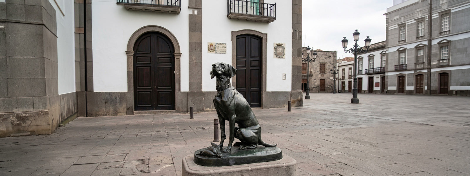 Bronze dog statue seated upright on a stone pedestal in a quiet European plaza with historic architecture, arched dark wooden doors, and ornate street lamps.