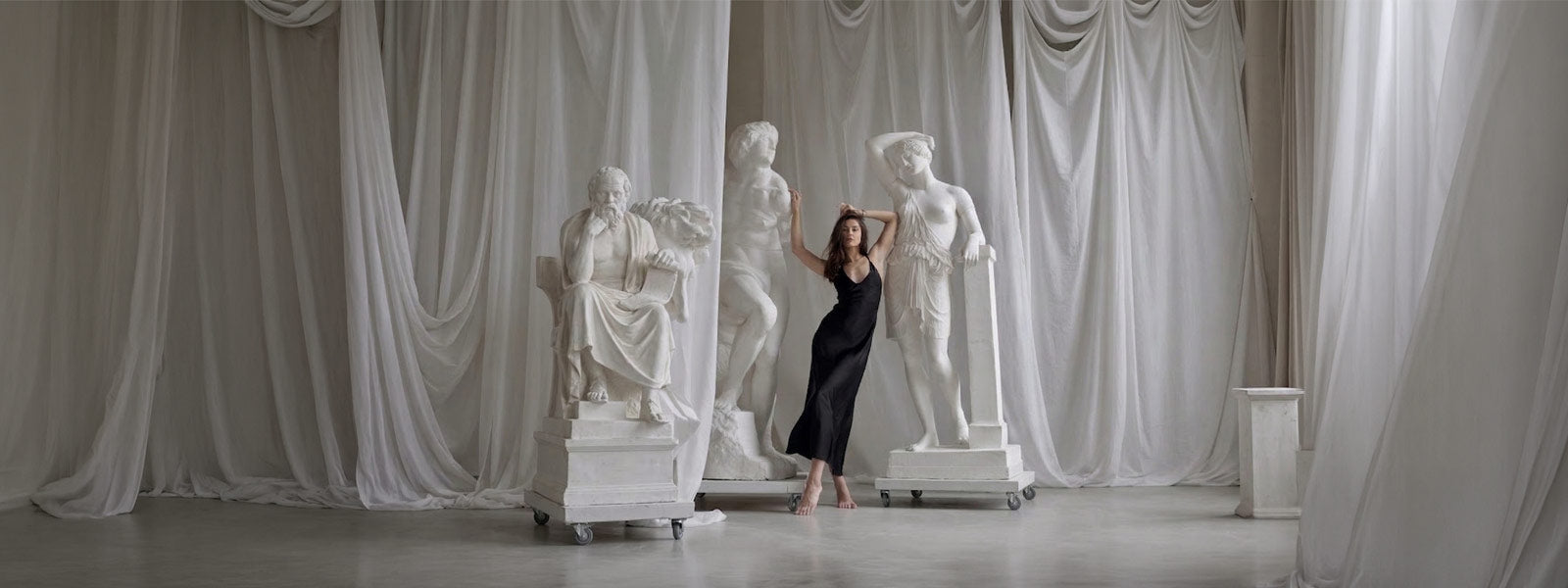 A barefoot woman in a black dress stands among three large white classical statues on wheeled platforms with flowing white curtains behind them.