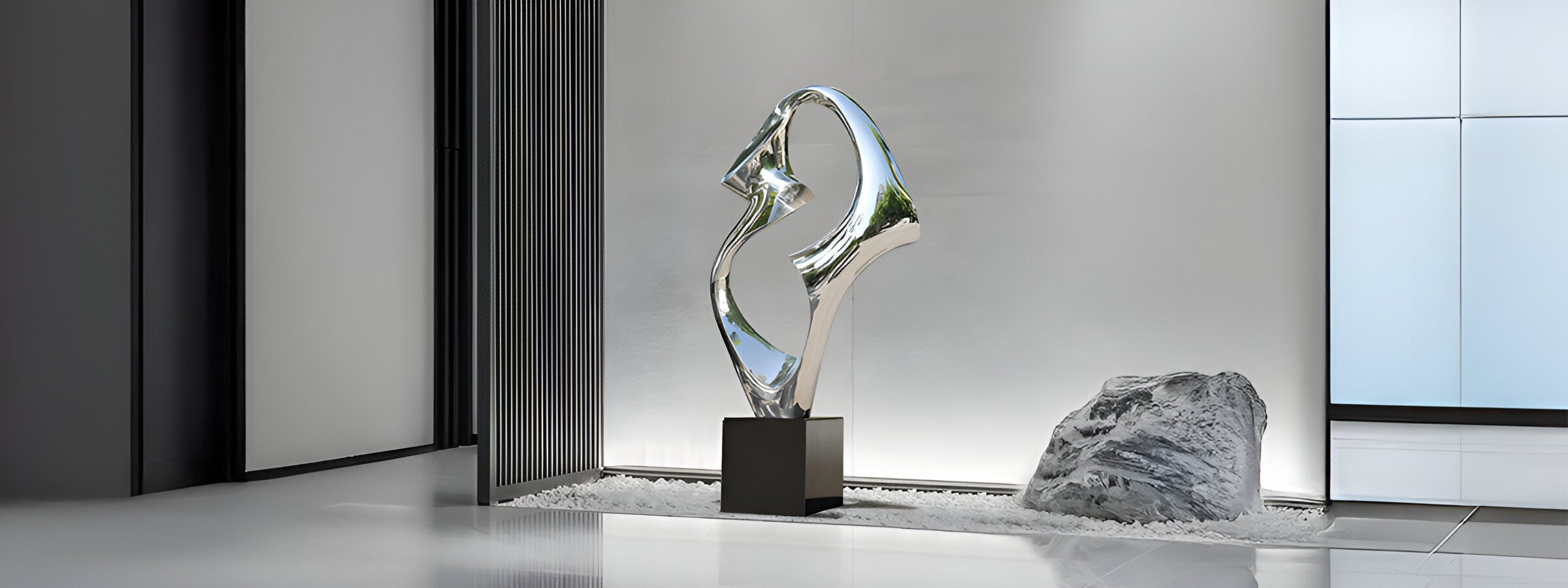 A modern sculpture with an abstract, metallic design stands on a circular platform. It is indoors, surrounded by marble walls, soft lighting, and a small leafless tree in a minimalist setting.