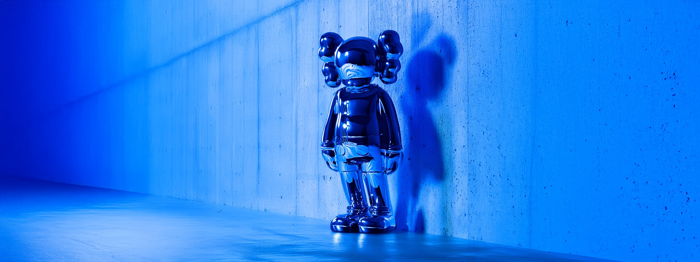 A shiny metallic sculpture of a humanoid figure with round ears stands against a blue-lit concrete wall, casting a shadow. The futuristic artwork reflects the ambient light, creating a captivating visual effect.