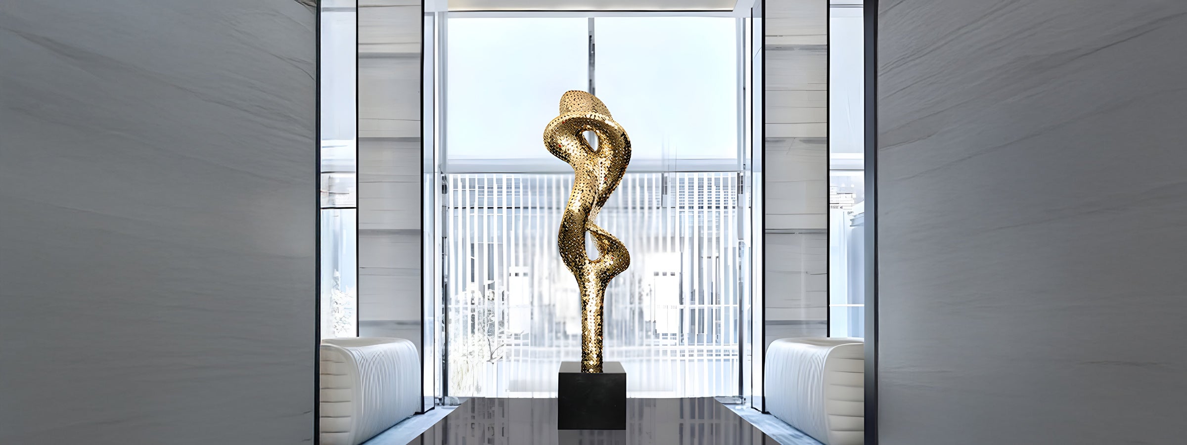 A tall, twisting golden sculpture stands on a black pedestal in a modern room with glass walls and white cushioned seating on either side. Natural light streams in through large windows in the background.