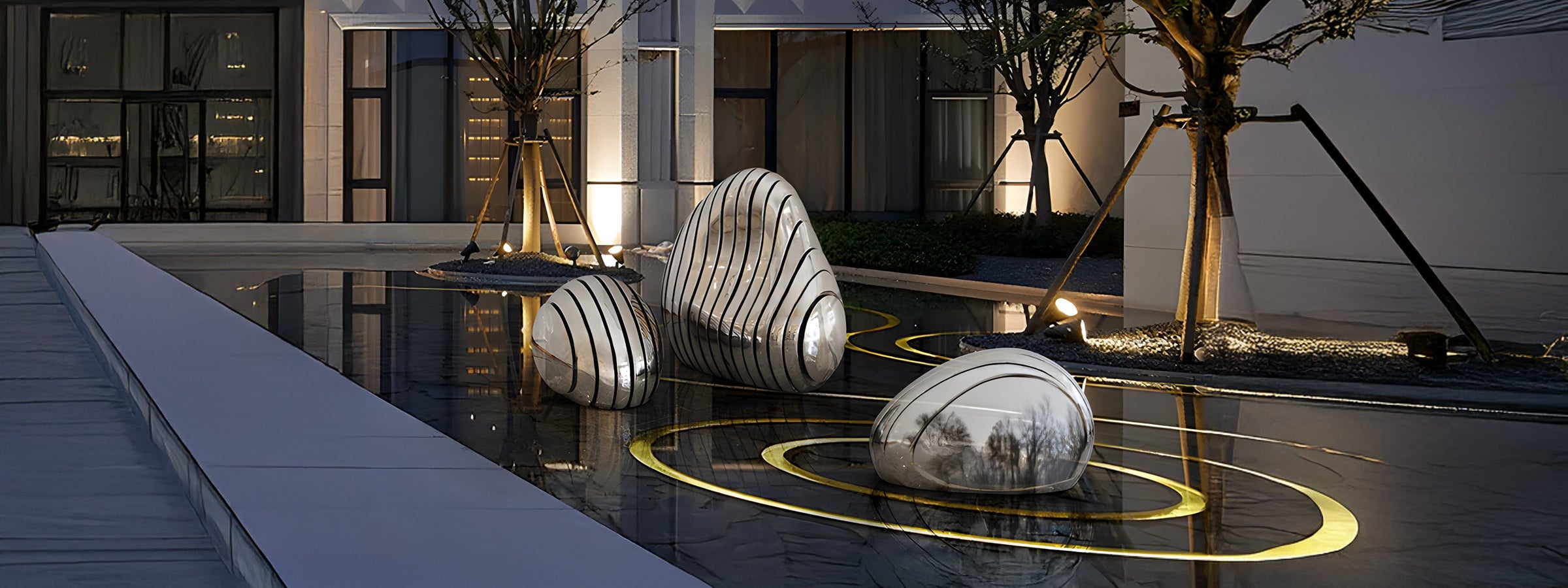 Three stainless steel pebble-shaped sculptures with black curved lines, reflecting the surrounding trees and lights. The sculptures are placed in a shallow reflective pool with circular lighting accents in a modern outdoor courtyard.