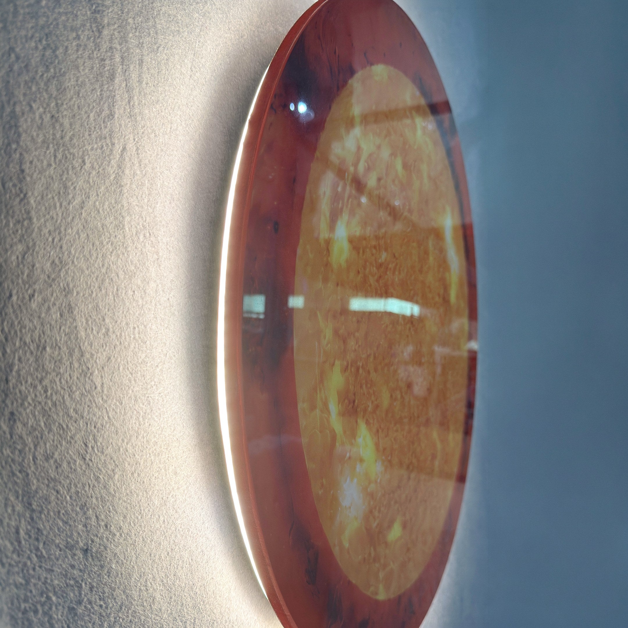 Side-angle view of the Galaxia Sun Crystal Porcelain LED Wall Art by Giant Sculptures, showing its amber and golden marbled porcelain surface with a brushed aluminum frame and warm LED backglow against a grey textured wall.