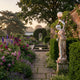 Giant Sculptures Dancing Lady Lamp Marble Sculpture 180cm in a walled garden, draped figure holding a globe lamp aloft beside a pond and floral path.