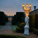 Classical Lady Five-Arm Lamp Marble Sculpture 300cm by Giant Sculptures, illuminated at dusk in a formal hedge-lined garden with an orangery beyond.
