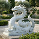 The Imperial Dragon White Marble Outdoor Sculpture - 120cm by Giant Sculptures stands on a stone platform in a landscaped garden, surrounded by hedges, flowering plants, and a reflecting pool.