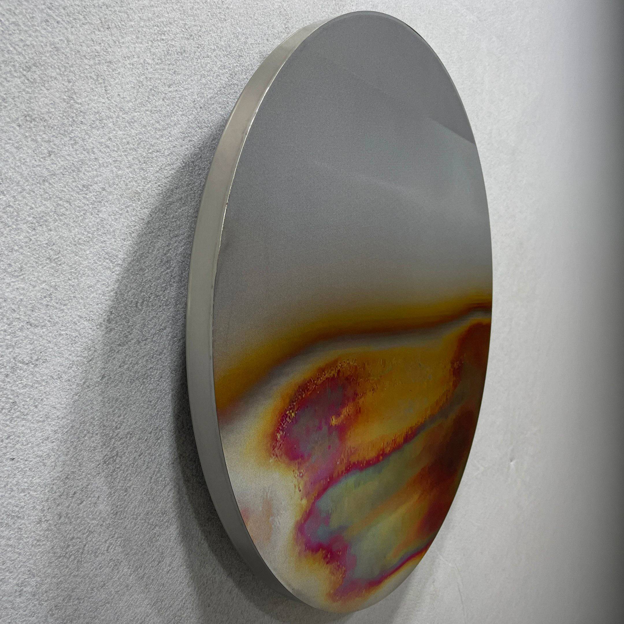 The Iridia Halo I Abstract Acrylic UV Wall Art by Giant Sculptures is a round metallic disc with a smooth surface, mounted on a light grey wall, featuring an iridescent blend of gold, red, pink, and blue shades near the bottom half.