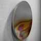 The Iridia Halo I Abstract Acrylic UV Wall Art by Giant Sculptures is a round metallic disc with a smooth surface, mounted on a light grey wall, featuring an iridescent blend of gold, red, pink, and blue shades near the bottom half.