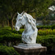 The Graceful Horse Head White Marble Outdoor Sculpture (60cm) by Giant Sculptures features a horse head with a flowing mane, set on a pedestal amidst lush greenery and trees.