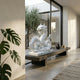 The Serpentine Dragon White Marble Outdoor Sculpture - 120cm by Giant Sculptures sits on a stone bench in a modern, minimalist room with large windows, potted plants, and views of the outdoor garden through the glass.