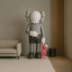 A modern design: The soft grey sculpture from Giant Sculptures features a gray cartoon-like figure with X eyes wearing gloves and black shorts, holding a smaller red figure. They stand beside a beige couch and black table, as part of the 180cm Soft Grey Iconify Pink Furry Toy Sculpture.
