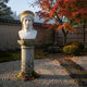 The Hera Marble Bust Sculpture (50cm) by Giant Sculptures stands on a pedestal in a serene garden with raked gravel, autumn leaves, and a vibrant Japanese maple. A stone path and bamboo fence complete the tranquil scene.