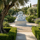 The Imperial Dragon White Marble Outdoor Sculpture - 120cm by Giant Sculptures stands on a pedestal amid manicured hedges and gravel paths, framed by an olive tree with a Mediterranean building behind.