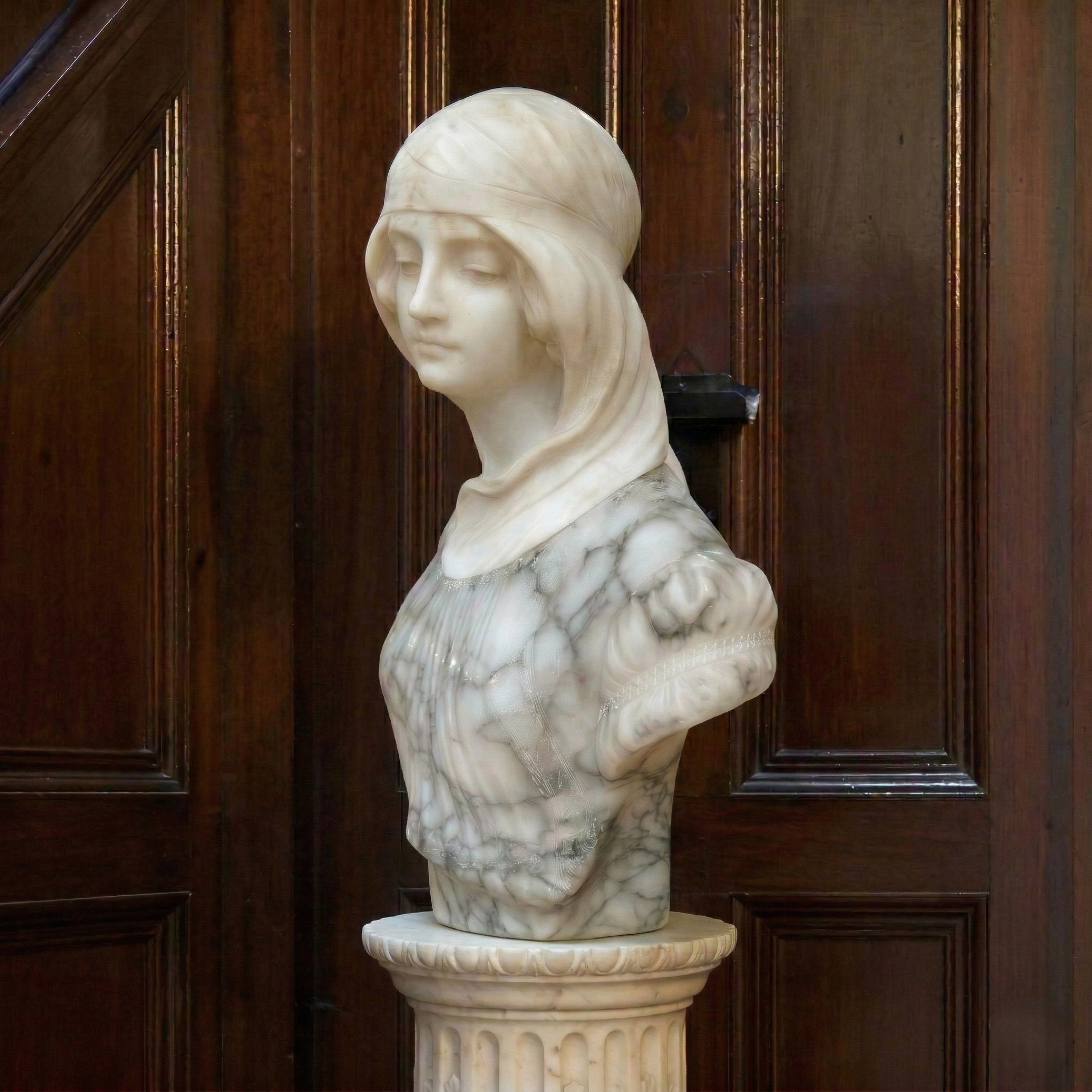 The Classical Maiden Marble Bust - 60cm by Giant Sculptures depicts a serene woman in a headscarf with downcast eyes, displayed on a decorative pedestal against dark wooden panels.