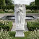 The Blooming Guardian Angel Marble Outdoor Sculpture - 220cm by Giant Sculptures stands in a manicured garden among roses and greenery, with a rectangular pond and stone pathway in the background.