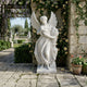 The 200cm Harvest Grace Angel Marble Outdoor Sculpture by Giant Sculptures stands on a stone patio among flowering vines, a potted plant, and lush greenery, with another angel marble outdoor sculpture in the background.