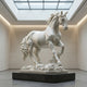 The Galloping Horse Marble Outdoor Sculpture - 160cm by Giant Sculptures stands as an impressive centerpiece in a modern gallery with white walls, polished floors, and a glass ceiling that fills the space with natural light.