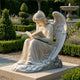 The Reflective Reading Angel Marble Outdoor Sculpture – 120cm by Giant Sculptures features a winged angel kneeling with an open book on a stone base, bringing serenity to any manicured garden with trimmed bushes, trees, and a water fountain backdrop.